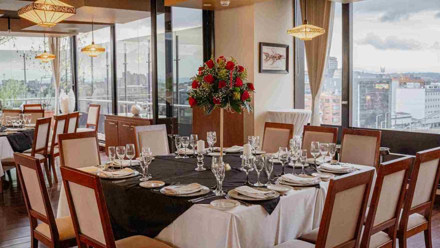 Un elegante comedor de restaurante con mesas puestas para un evento, decorado con un gran arreglo floral y vistas a la ciudad.