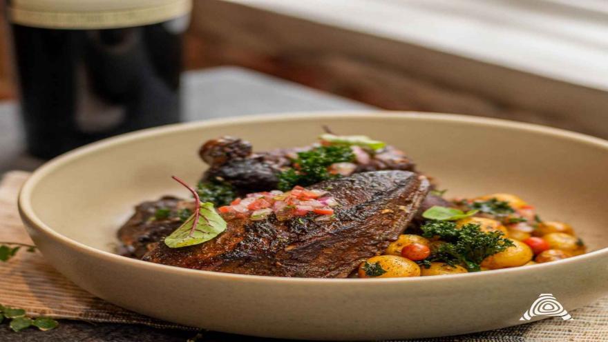 Un plato elegante con un corte de carne oscura, acompañado de papas baby y una fresca ensalada picada, en un ambiente acogedor.
