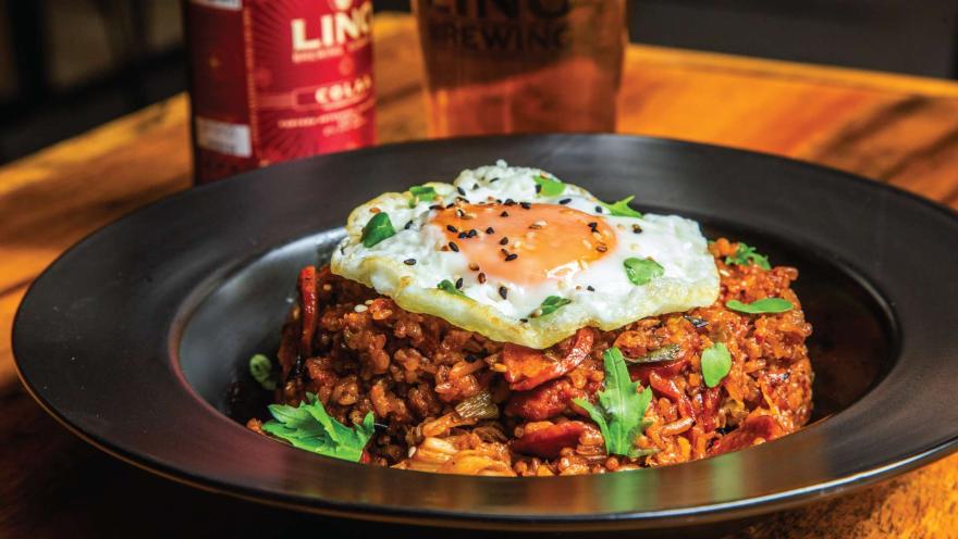 Un plato de arroz frito picante con kimchi, coronado con un huevo frito y espolvoreado con semillas de sésamo, con una bebida de fondo.