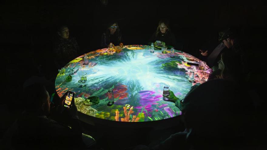  Comensales alrededor de una mesa redonda con proyecciones interactivas de vida marina, en un ambiente oscuro.