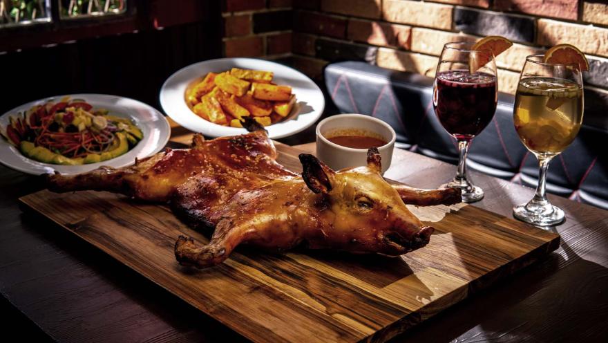 Un cochinillo asado entero sobre una tabla de madera, acompañado de guarniciones y copas de vino.