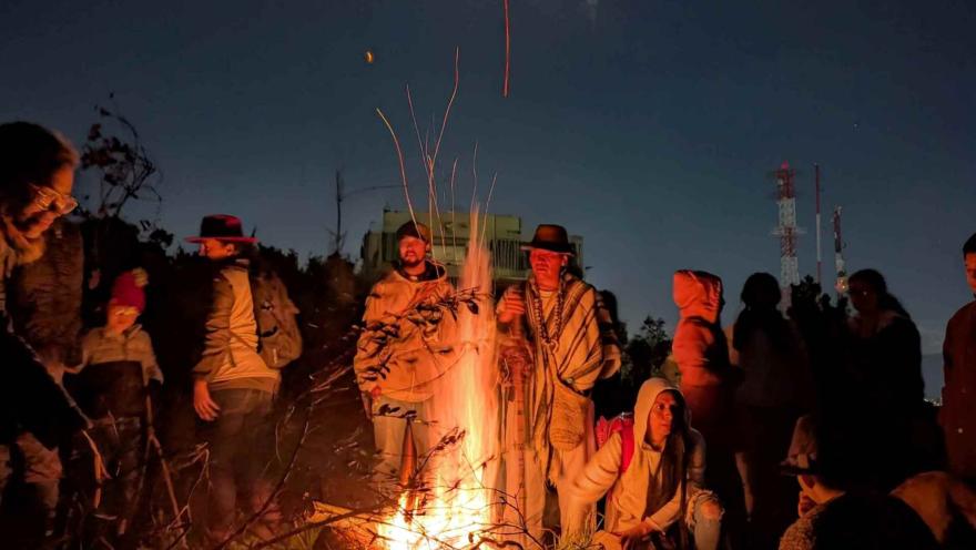 Grupo de personas entre jóvenes y adultos de noche, con chaquetas, reunidos alrededor de una chimenea. En el centro está una persona que parece un campesino o indigena con sombrero y ruana.