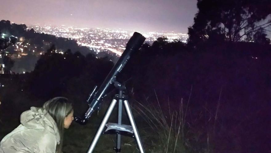 Mujer joven con una chaqueta clara observa de noche a través de un telescopio dirigido hacia las estrellas.