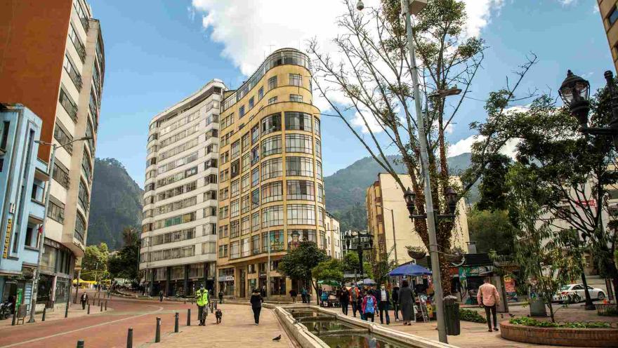 A promenade with a long fountain and an old building with many windows on one corner. There are trees, a security guard with a dog, and people nearby.