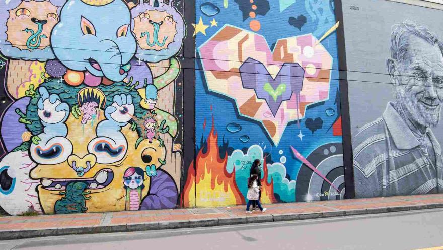 Calle en la que se observan tres paredes pintadas con graffitis. En una hay un elefante psicodélico; en otra corazones de colores; y en otra en blanco y negro un rostro de un hombre anciano. Una mujer y una niña caminan por el frente.