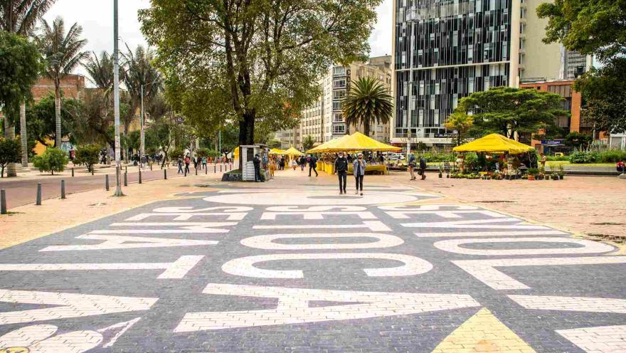 A street with signs painted on the sidewalk and people walking along it. Some yellow awnings and a booth with a security guard can be seen in the background.