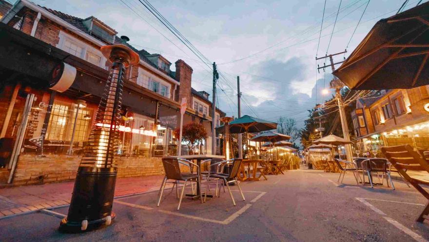  Una calle al atardecer con mesas alrededor cubiertas por carpas. Cada una tiene un calentador de ambiente de gas en forma de pirámide.