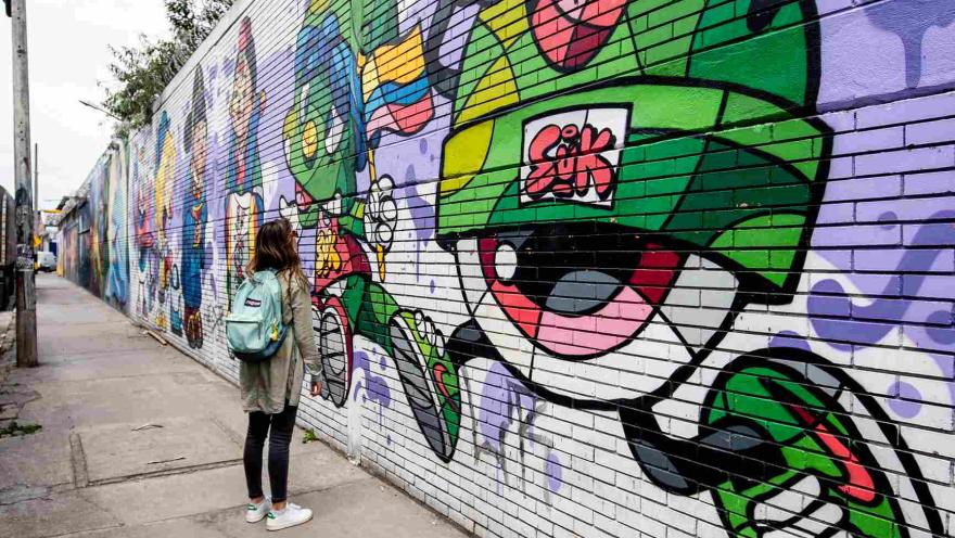 Mujer joven observa una pared en la que hay un graffiti con figuras coloridas.