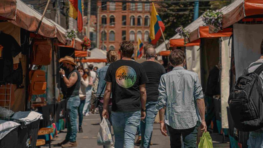 Hombres jóvenes, parecen turistas, que caminan al frente de unos toldos de comercio callejero. En los lugares se observan varias banderas de Colombia.