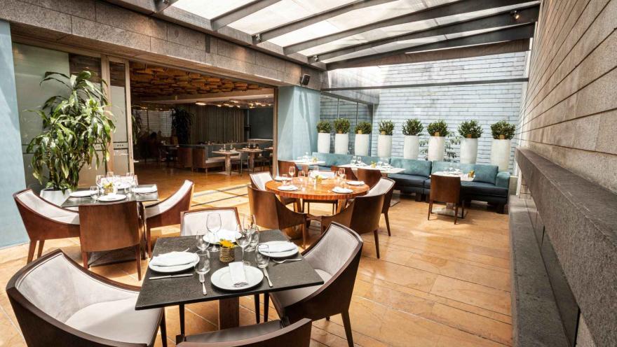 Un restaurante moderno con una sección interior y una terraza cubierta, mesas elegantes y cómodos sofás. La decoración es sofisticada, con plantas y tonos neutros.