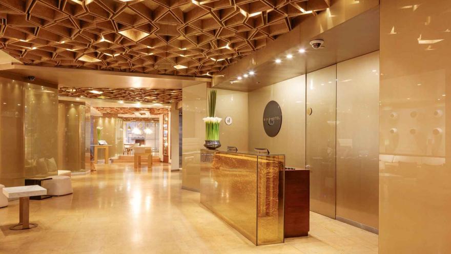  Un elegante lobby de hotel con una recepción dorada brillante, un techo con diseño geométrico y una decoración minimalista. El ambiente es lujoso y acogedor.