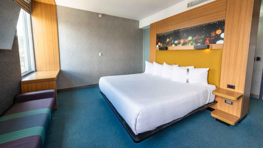 Una habitación de hotel Aloft moderna y colorida con una cama king-size, una ventana con banco y un cabecero decorativo con luces.