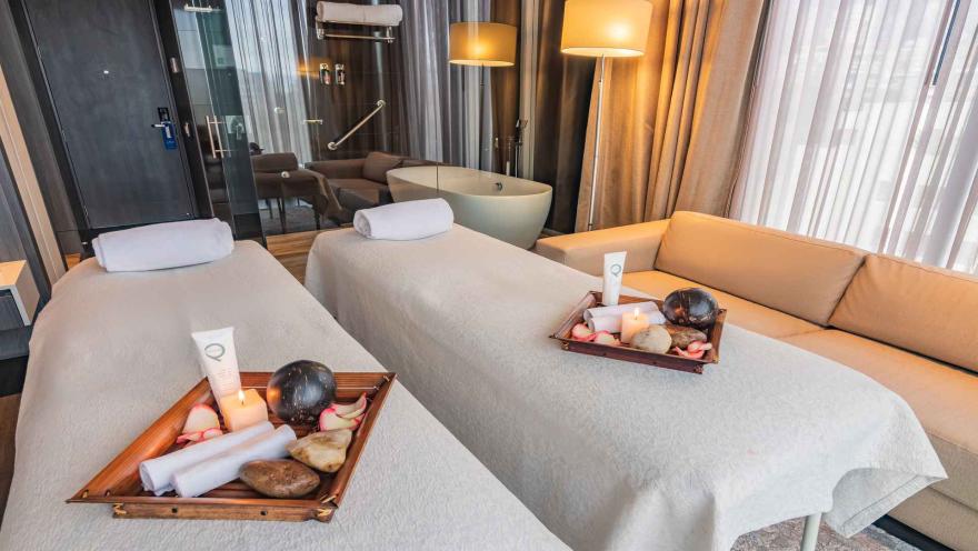 Dos camillas de masaje preparadas en una suite de hotel con una bañera independiente al fondo. El ambiente es lujoso y relajante, ideal para tratamientos de spa privados.