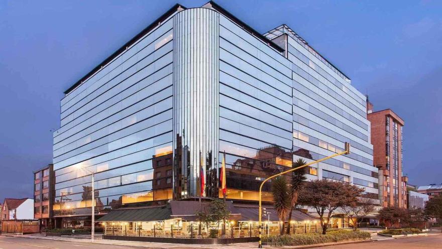  Un moderno edificio de hotel con fachada de cristal, que refleja el entorno urbano al atardecer o al anochecer. La iluminación del restaurante en la planta baja añade calidez.