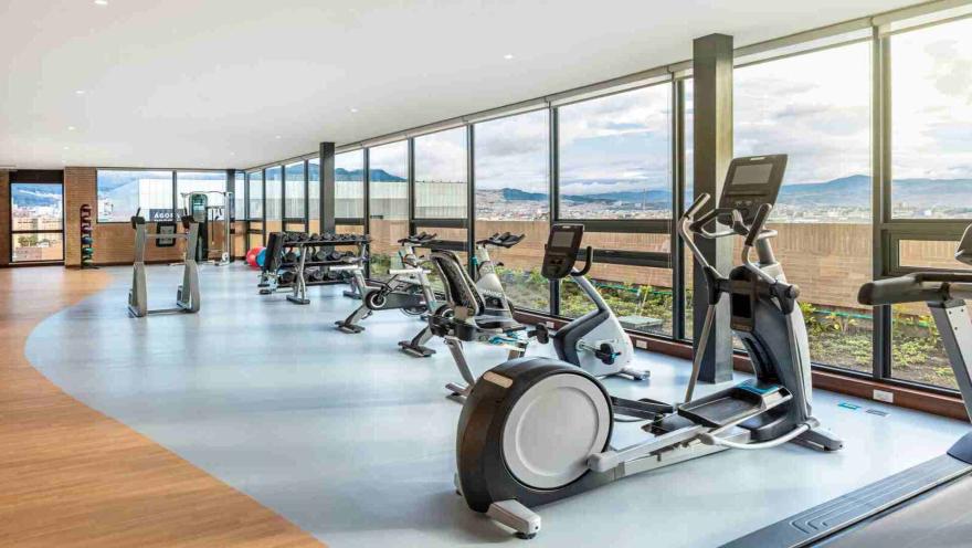 Un gimnasio espacioso y bien iluminado con grandes ventanales que ofrecen vistas panorámicas de la ciudad y las montañas. Equipado con diversas máquinas de ejercicio.