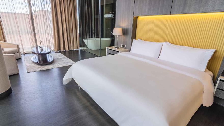 Una lujosa habitación de hotel con una cama king-size, cabecera amarilla, y un baño con bañera independiente visible a través de un panel de cristal.