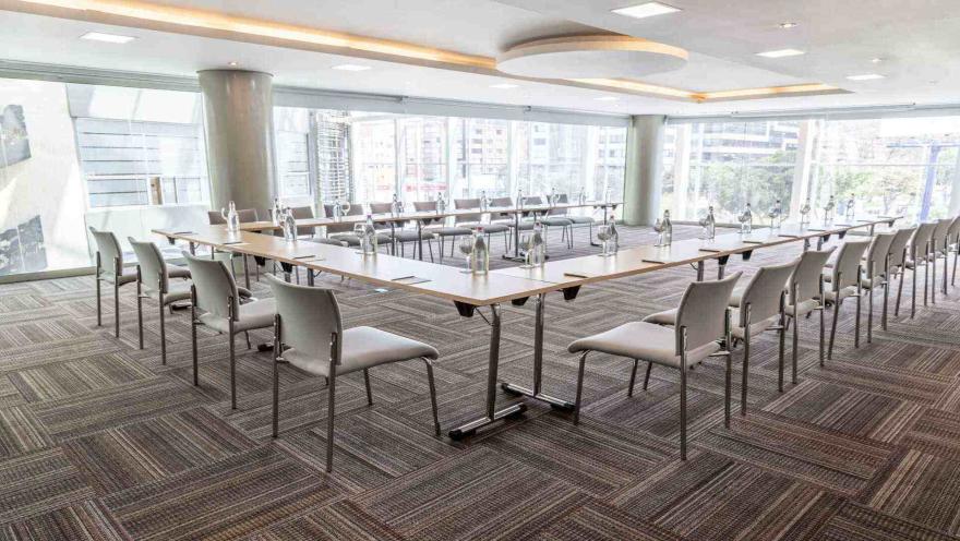  Una sala de reuniones dispuesta en forma de "U" con mesas y sillas. Grandes ventanales ofrecen vistas de la ciudad, creando un ambiente de trabajo dinámico.