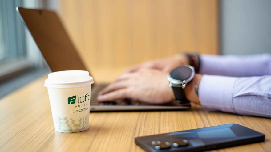 Un primer plano de una taza de café "Aloft Hotels" en un escritorio, con las manos de una persona trabajando en un portátil y un smartphone de fondo.