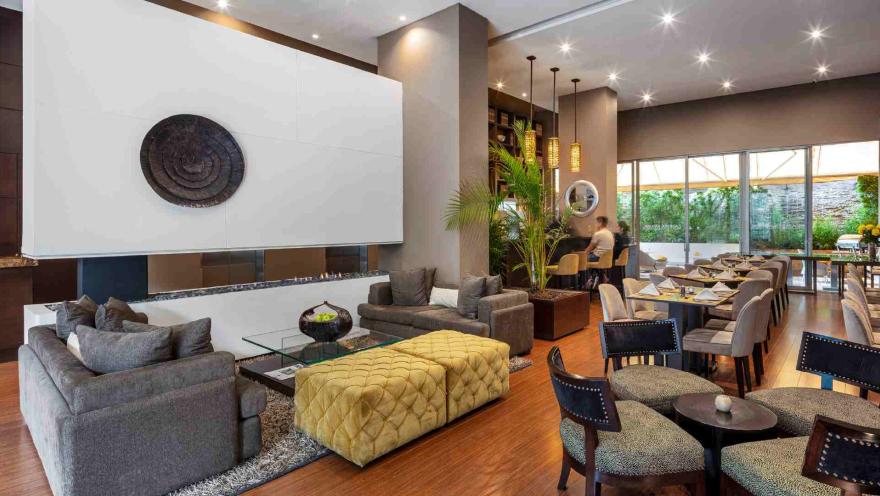 Un moderno lobby lounge que combina un área de sofás frente a una chimenea y un comedor. La decoración es sofisticada, con toques de verde y madera.