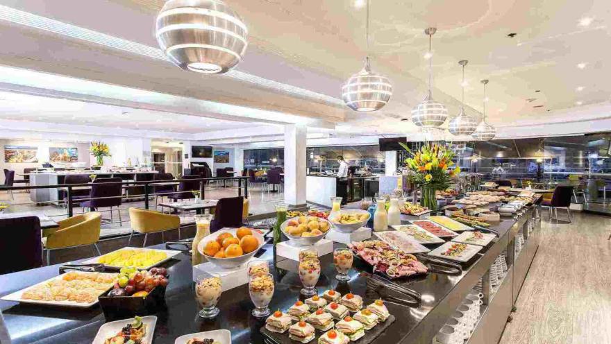 Un extenso buffet con una gran variedad de alimentos, incluyendo frutas frescas, pasteles, quesos y embutidos, presentado de manera atractiva en un ambiente elegante.
