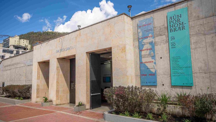 Fachada de edificación en piedra en la que destacan un par de pendones y una leyenda sobre la parte superior que dice: “GALERÍA SANTA FE”. Al fondo se observa el cerro de Monserrate. 