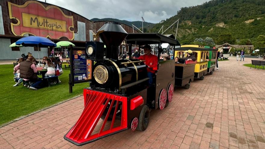 Un tren en miniatura en un parque infantil. Hay personas alrededor y al fondo se observa una montaña.