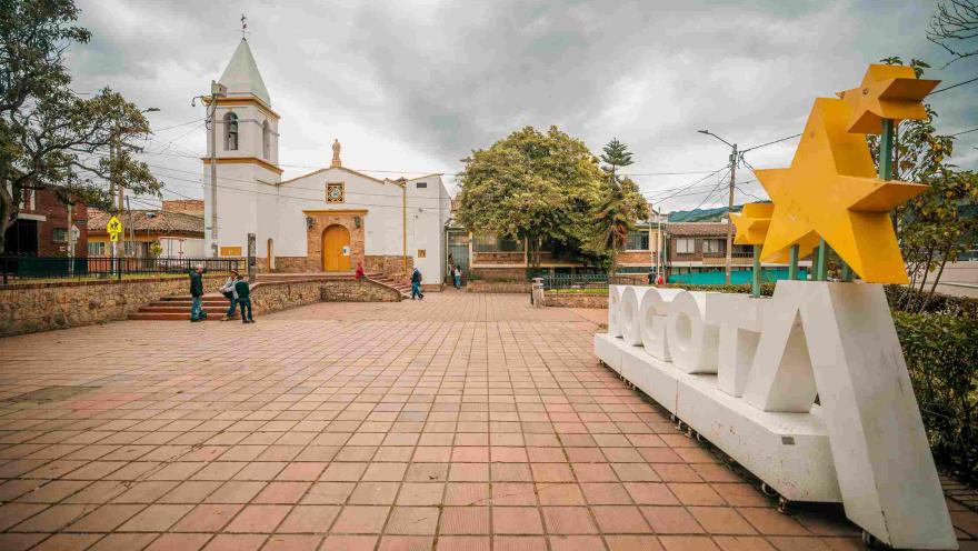 Wide shot of the square featuring a white church with yellow accents in the background. On the right, there is a large white sign with the word "Bogotá", featuring yellow stars above the letter “A.”