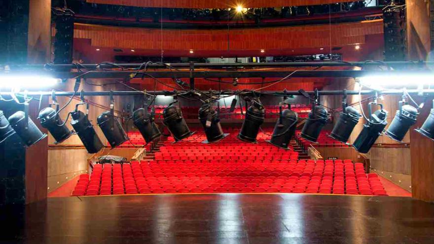 Primer plano de focos de luz en un teatro. Al fondo se observan las sillas en el escenario.