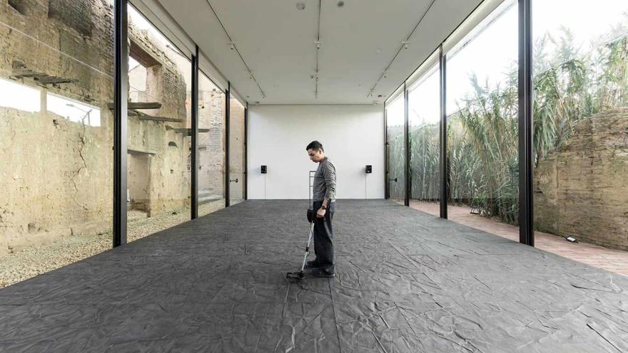 Hombre joven de pie en un salón sobre un piso, que parece ser de baldosa de color negro, mira al suelo y lo toca con un aparato que parece un detector de metáles. 