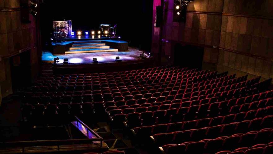 Plano general de un teatro en el que con poca iluminación se observan la silletería y la tarima, en la que hay algunas luces.