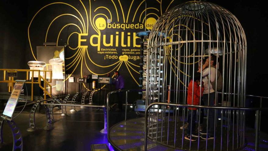 Una jaula gigante metálica en cuyo interior hay un hombre joven y un niño tapandose los oidos con las manos.