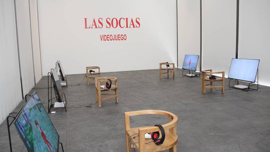 Un salón con varias pantallas de televisión sobre el suelo con una silla al frente cada una y en ellas un par de audífonos grandes. Hay una pared gris al frente con una leyenda en letras rojas que dice: “Las Socias Videojuego”.