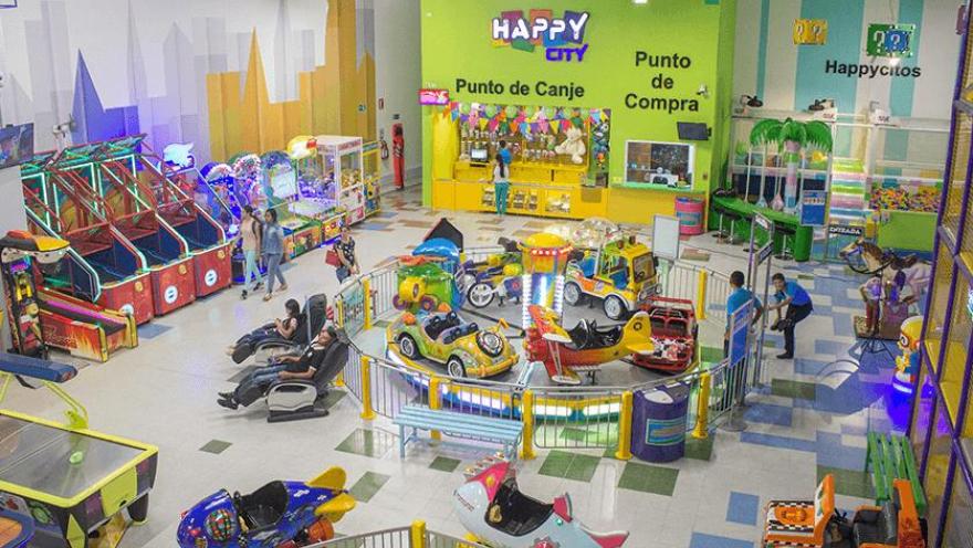 Salón grande en el que hay varias atraccciones mecánicas con coloridas figuras de animales y cosas. Hay varias personas en el lugar.