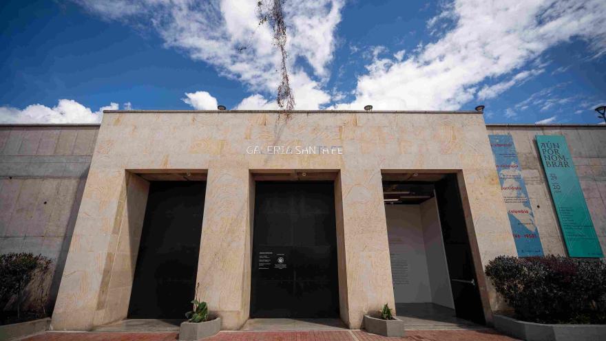 Fachada de edificación en piedra en la que destacan tres puertas negras y altas, parece que de metal, una de ellas abierta y una leyenda sobre la parte superior que dice: “GALERÍA SANTA FE”. 