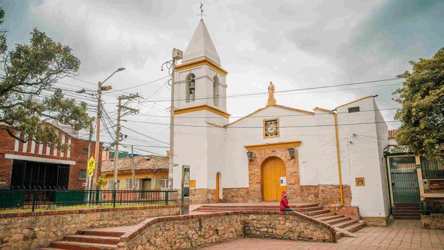 Close-up of a white church with yellow accents. In front of it, there is a person wearing a red jacket.