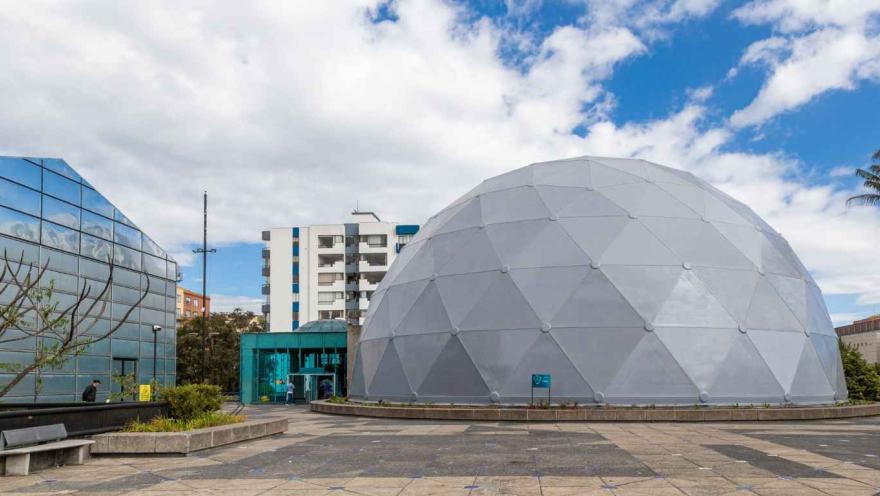 Estructura en forma de domo de color gris, rodeada por un edificio moderno y una estructura de vidrio. 