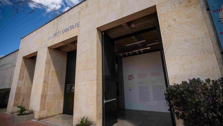 Fachada de edificación en piedra en la que destacan tres puertas negras y altas, parece que de metal, una de ellas abierta y una leyenda sobre la parte superior que dice: “GALERÍA SANTA FE”. Hay tres macetas con plantas pequeñas alrededor de la estructura.