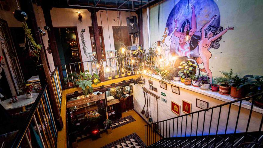 Salón interior de una casa en la que resaltan escaleras y balcones. Hay una pared con un dibujo de dos mujeres desnudas bailando, hay varias materas con plantas, y varias lámparas encendidas que cuelgan del techo.