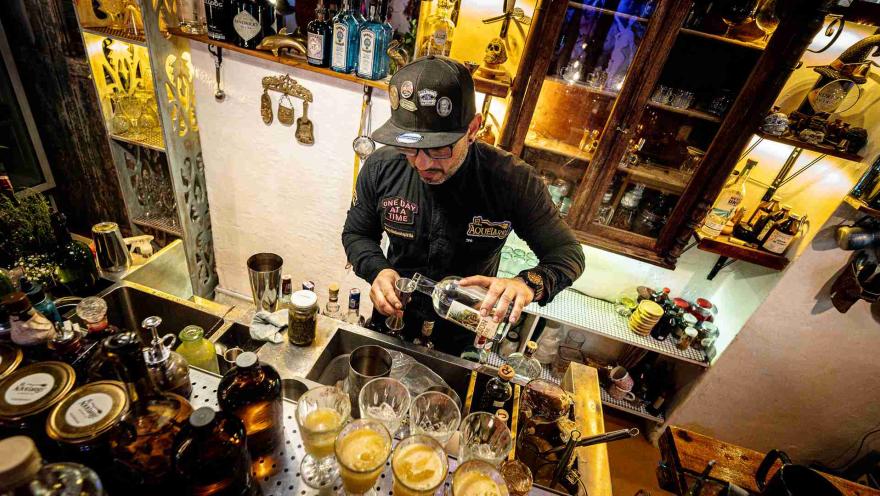 Hombre joven sirviendo algo parecido a alcohol en copas de cristal y metal.  Detrás del hombre en una pared hay muchos objetos de decoración.