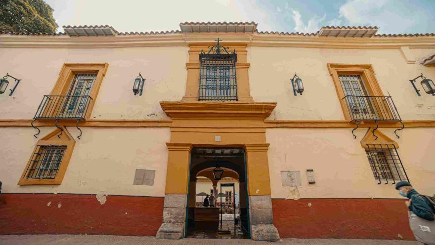 Fachada de casa antigua en la que se destacan sus colores crema, amarillo y rojo, y sus dos ventanas con rejas en hierro.