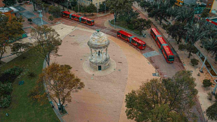 Plazoleta vista desde arriba en la que sobresale un templete hecho en piedra y rodeado de vegetación. Algunos buses rojos de TransMilenio transitan alrededor