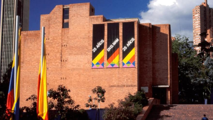 Fachada de gran edificio en ladrillo donde sobresalen avisos con una leyenda en letras azules que dicen: “25 AÑOS EN EL MAM”. A un lado hay una bandera de Colombia y otra de Bogotá.