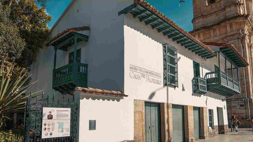 Vista de lado de la fachada de una casa antigua de estilo colonial con balcones y puertas de color verde. Resalta una leyenda en una de las paredes blancas, que dice: “Museo de la Independencia - Casa del Florero”.