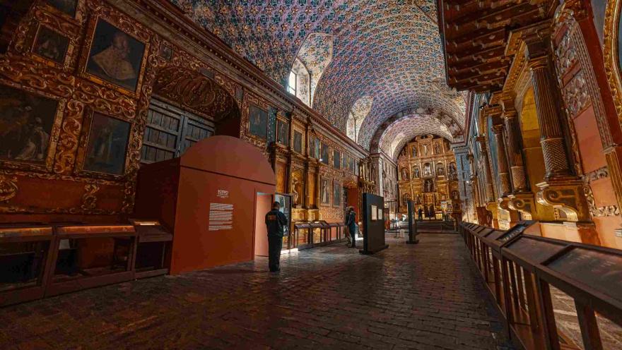 Interior de un recinto religioso, adornado por varios cuadros y retablos. El techo está adornado con unas flores azules y doradas.