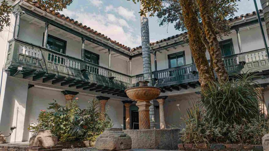 Patio interior de casa antigua en la que sobresalen unos balcones en el segundo piso y una fuente de agua en el centro, rodeada por árboles y otra vegetación.
