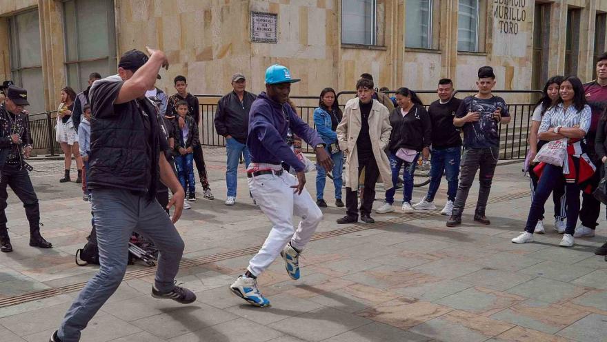 Un par de hombres jóvenes bailan pasos modernos en una calle; hay personas alrededor observándolos.