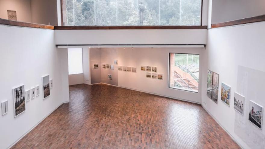 Sala interior de dos pisos bien iluminada con un ventanal grande hacia la calle y varios cuadros en las paredes pintadas de blanco.