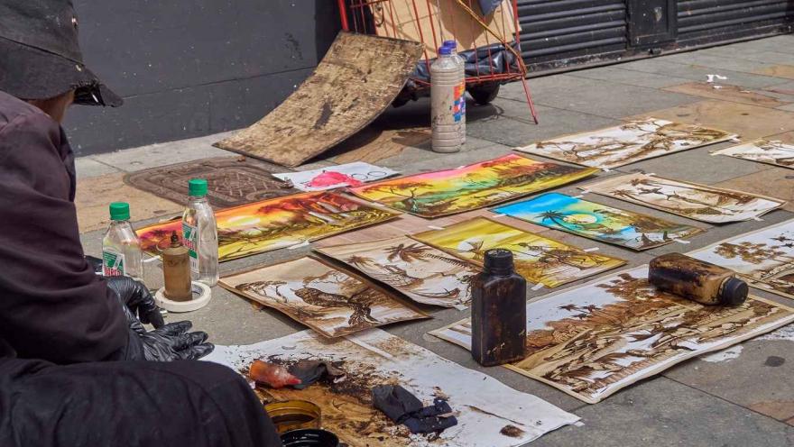 Una suerte de pinturas, parece que recién hechas, expuestas sobre el piso de una calle. Se observa de un lado al artista de las obras.