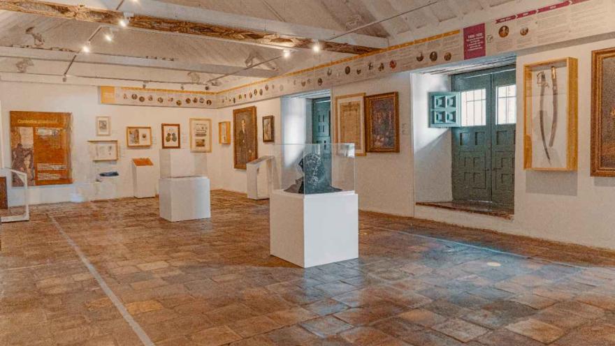 Salón interior con varias piezas en exhibición en las paredes y en el suelo dentro de unas vitrinas. Hay una ventana un poco abierta y de la cual entra algo de luz natural.