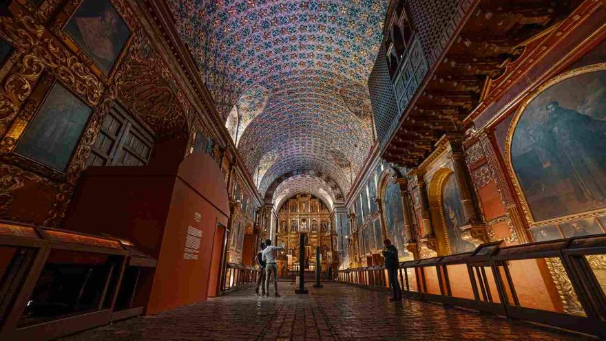  Interior de un recinto religioso, adornado por varios cuadros y retablos. Hay tre hombres que observan el techo y las paredes del lugar.